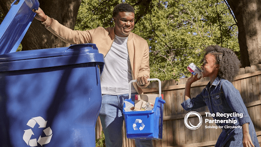 A father and daughter participating in recycling together outdoors. The father lifts the lid of a large blue recycling bin while the daughter holds a smaller blue recycling basket and drinks from a recyclable can. The Recycling Partnership logo and tagline 'Solving for Circularity' appear in the bottom right corner.
